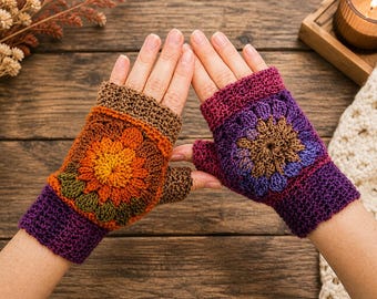 Op de afbeelding: Een paar handgemaakte vingerloze handschoenen in tinten oranje, bruin, paars en groen. De handschoenen hebben een bloemmotief op de rug van de hand en zijn gemaakt van een gehaakt materiaal. De handschoenen worden op een houten oppervlak gepresenteerd.