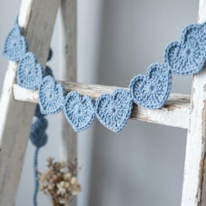 May include: A garland featuring a series of light blue crocheted hearts connected by a matching yarn string. Each heart is crafted with a detailed circular stitch pattern in the center, radiating outward to form the heart shape, creating a continuous chain.