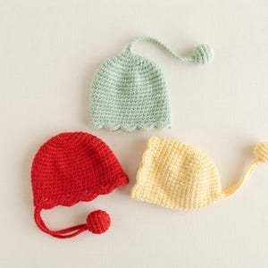 May include: Three crocheted hats in light green, bright red, and pale yellow. Each hat features a scalloped edge and a long, thin cord ending in a small, round crocheted ball. The hats are crafted with a consistent, textured yarn stitch pattern.