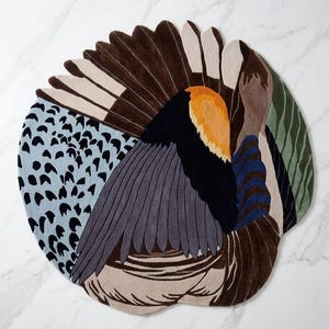 K&ouml;nnte beinhalten: Ein runder Teppich mit einem stilisierten Vogelmotiv und gef&auml;chertem Schwanz. Das Muster besteht aus hellblauen Bereichen mit schwarzen Punkten, Dunkelbraun, Grau, einem leuchtend orangefarbenen Fleck, dunkelblauen Streifen und gr&uuml;nen Wellenlinien.
