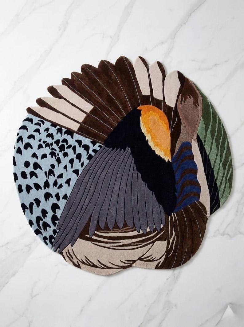K&ouml;nnte beinhalten: Ein runder Teppich mit einem stilisierten Vogelmotiv und gef&auml;chertem Schwanz. Das Muster besteht aus hellblauen Bereichen mit schwarzen Punkten, Dunkelbraun, Grau, einem leuchtend orangefarbenen Fleck, dunkelblauen Streifen und gr&uuml;nen Wellenlinien.