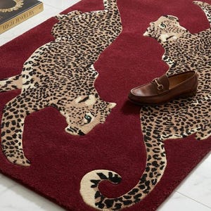 May include: A rectangular deep red rug featuring two large, beige and black leopard figures with spotted patterns. The rug has a soft, tufted texture and a small metallic rectangular tag at the bottom edge. A brown leather loafer rests on the rug.