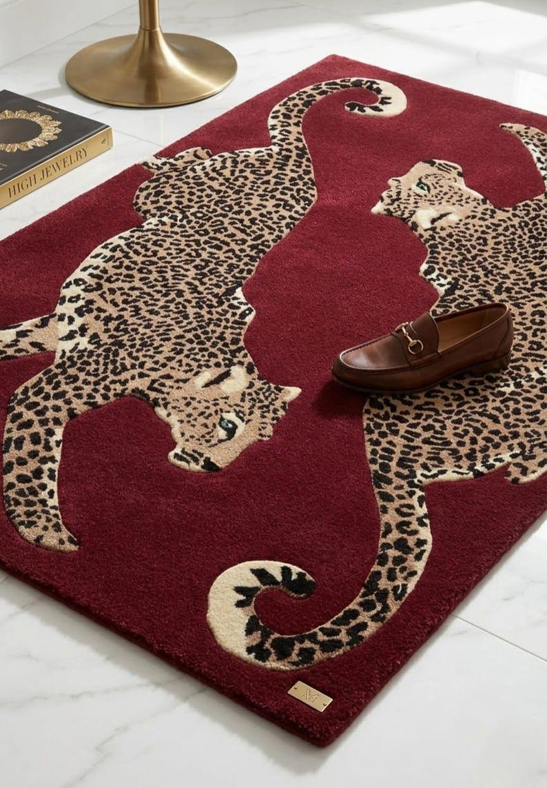 May include: A rectangular deep red rug featuring two large, beige and black leopard figures with spotted patterns. The rug has a soft, tufted texture and a small metallic rectangular tag at the bottom edge. A brown leather loafer rests on the rug.