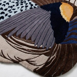 K&ouml;nnte beinhalten: Ein getufteter Teppich mit einem abstrakten Vogelfl&uuml;gel-Design in Braun-, Grau-, Dunkelblau- und Gelbt&ouml;nen. Die Oberfl&auml;che zeigt strukturierte Muster, darunter einfarbige Bl&ouml;cke, geschichtete federartige Formen und einen hellblauen Bereich mit schwarzen Punkten.
