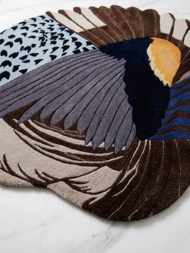 K&ouml;nnte beinhalten: Ein getufteter Teppich mit einem abstrakten Vogelfl&uuml;gel-Design in Braun-, Grau-, Dunkelblau- und Gelbt&ouml;nen. Die Oberfl&auml;che zeigt strukturierte Muster, darunter einfarbige Bl&ouml;cke, geschichtete federartige Formen und einen hellblauen Bereich mit schwarzen Punkten.