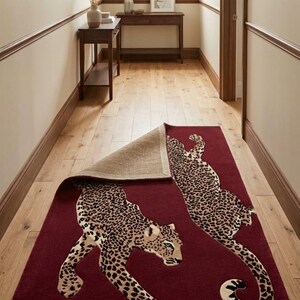 May include: A rectangular area rug in a deep burgundy red color featuring two large leopard figures with black and tan spots. One corner of the rug is folded back to reveal a textured, light brown woven backing material underneath the pile.