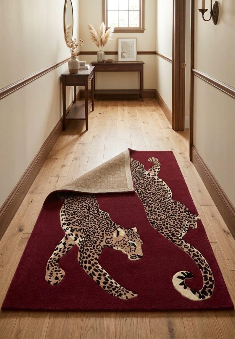 May include: A rectangular area rug in a deep burgundy red color featuring two large leopard figures with black and tan spots. One corner of the rug is folded back to reveal a textured, light brown woven backing material underneath the pile.
