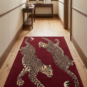 May include: A rectangular area rug in a deep burgundy red color featuring two large, stylized leopard figures in a tan and black spotted pattern. The leopards are positioned in a dynamic, leaping pose across the center of the rug surface.