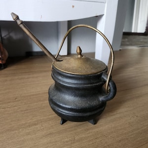 May include: A black cast iron pot with three small feet, a rounded body, and a side handle. It features a brass-colored lid with a central finial and a long, thin, angled brass-colored spout extending from the top opening of the pot.