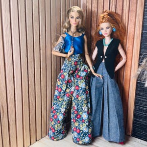 May include: Two fashion dolls standing side by side. The doll on the left wears a blue sleeveless top, floral print wide-leg trousers, and a silver handbag. The doll on the right wears a black sleeveless vest, a turquoise necklace, and grey trousers.