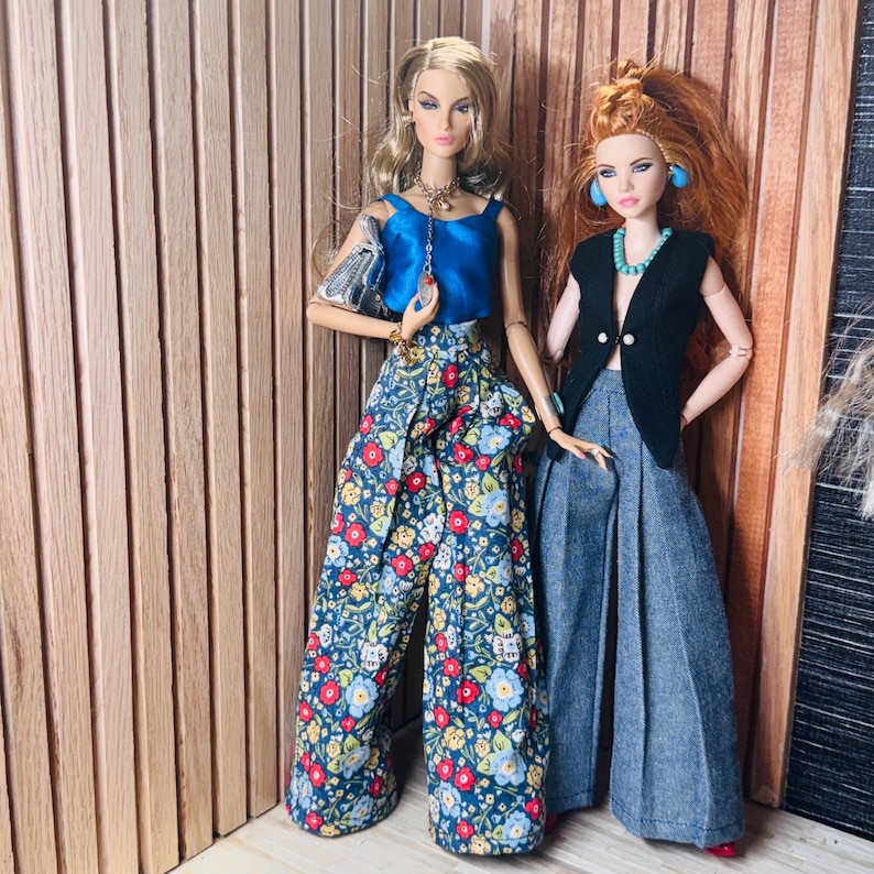 May include: Two fashion dolls standing side by side. The doll on the left wears a blue sleeveless top, floral print wide-leg trousers, and a silver handbag. The doll on the right wears a black sleeveless vest, a turquoise necklace, and grey trousers.
