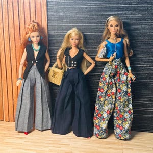 May include: Three fashion dolls standing side by side. The first has red hair, a black vest, and gray wide-leg trousers. The second has blonde hair, a black cropped vest, dark trousers, and a tan handbag. The third has blonde hair, a blue top, and floral trousers.