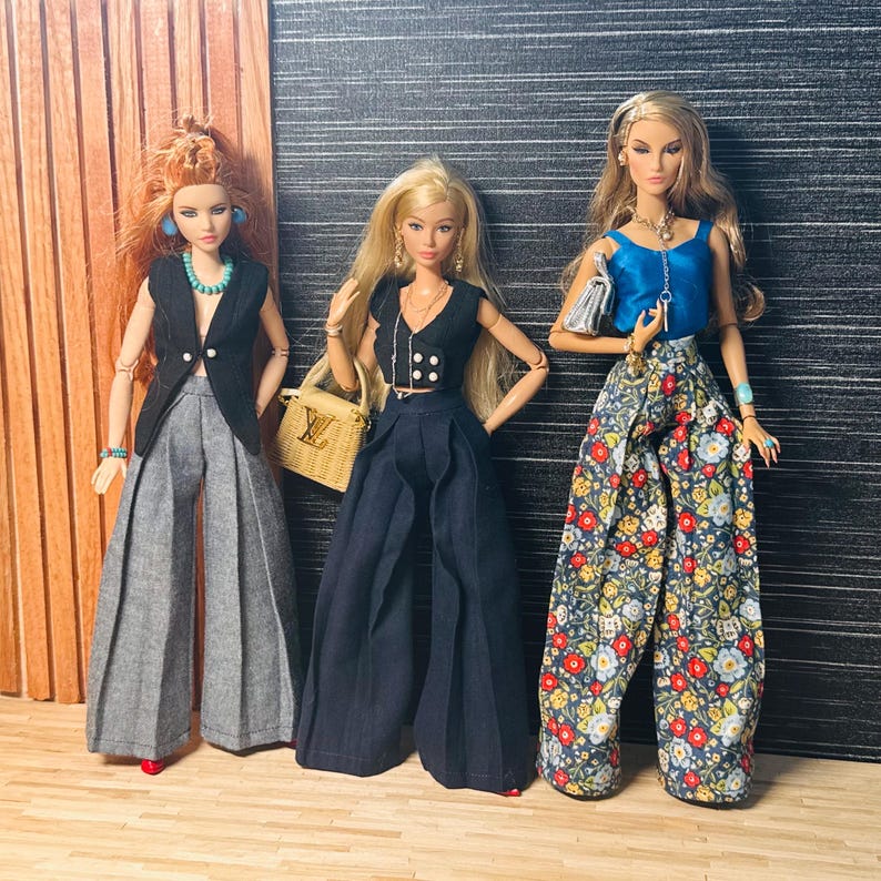 May include: Three fashion dolls standing side by side. The first has red hair, a black vest, and gray wide-leg trousers. The second has blonde hair, a black cropped vest, dark trousers, and a tan handbag. The third has blonde hair, a blue top, and floral trousers.