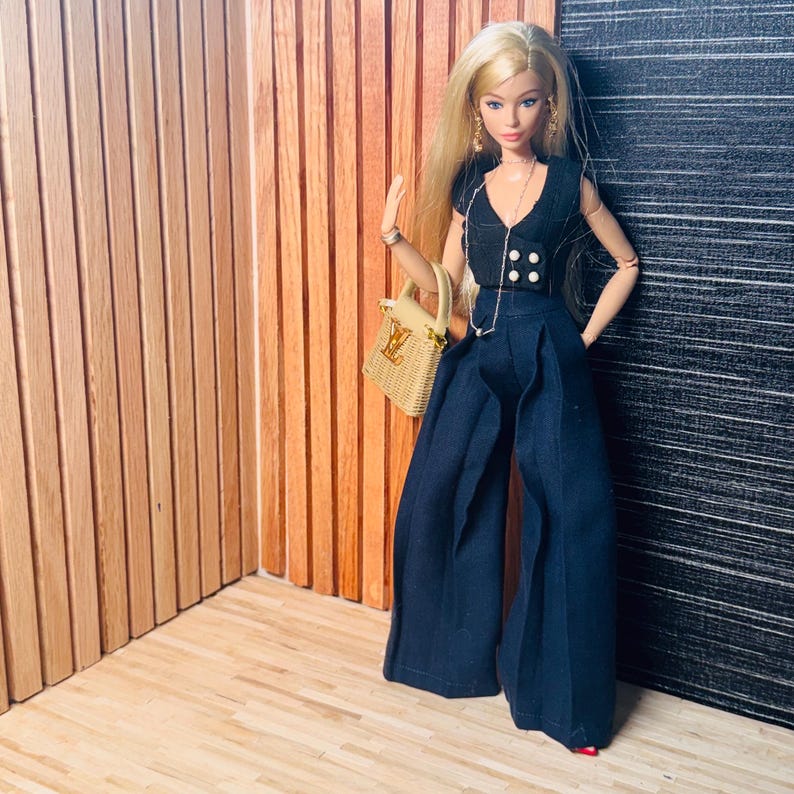 May include: A blonde doll wearing a black sleeveless vest with white buttons and matching wide-leg trousers. The doll features a long silver chain necklace with a small pendant and carries a tan woven handbag with a gold-toned logo emblem.