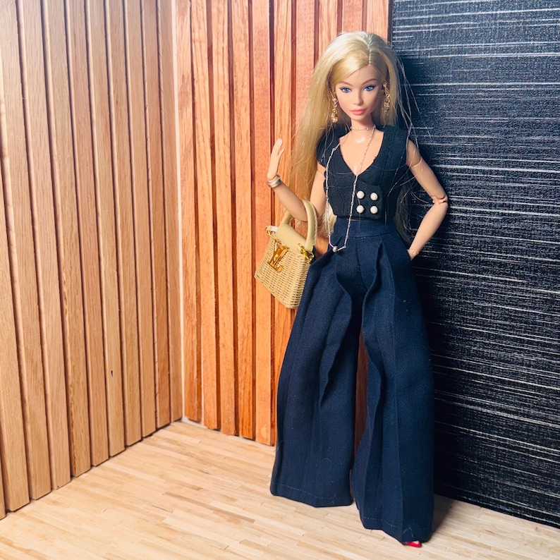 May include: A blonde doll wearing a black sleeveless top with four white buttons and wide-leg black trousers. The doll features gold dangling earrings, a thin silver necklace, and carries a small tan woven handbag with a gold logo emblem.