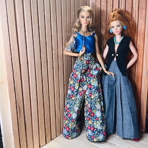May include: Two fashion dolls standing side by side. The left doll wears a blue satin sleeveless top, floral print wide-leg trousers, and a silver handbag. The right doll wears a black sleeveless vest, a turquoise necklace, and grey wide-leg trousers.