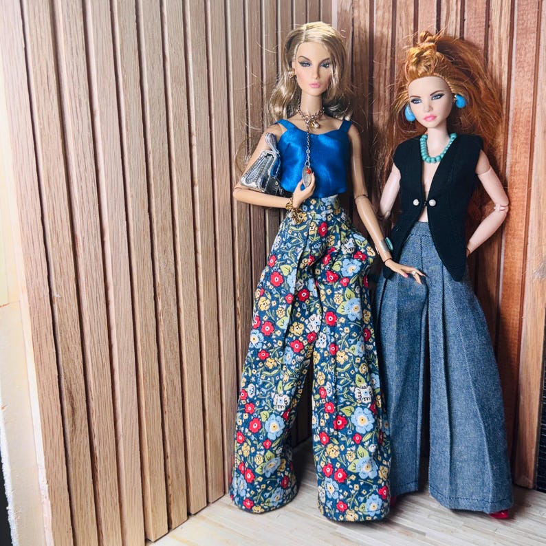 May include: Two fashion dolls standing side by side. The left doll wears a blue satin sleeveless top, floral print wide-leg trousers, and a silver handbag. The right doll wears a black sleeveless vest, a turquoise necklace, and grey wide-leg trousers.