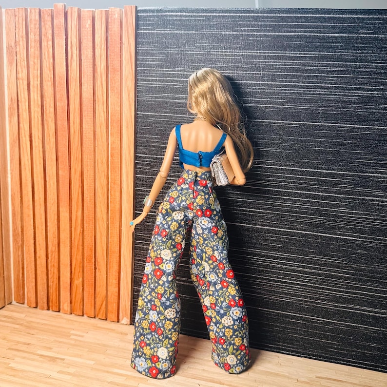 May include: A doll with long blonde hair wearing a bright blue sleeveless crop top and high-waisted wide-leg trousers featuring a dark blue background with a colorful floral pattern of red, white, and yellow flowers. The doll carries a small beige clutch.