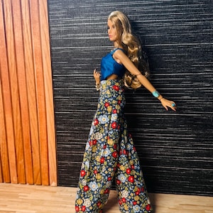 May include: A doll with long blonde hair wearing a sleeveless blue top and wide-leg trousers featuring a dark blue background with a multi-colored floral pattern of red, yellow, and light blue flowers. The doll wears a turquoise bracelet on one wrist.