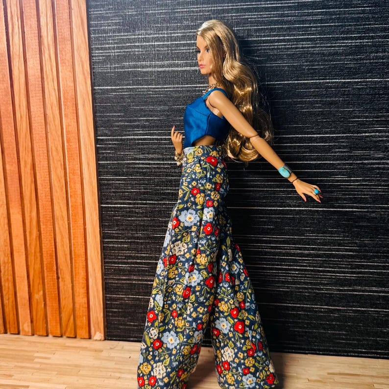 May include: A doll with long blonde hair wearing a sleeveless blue top and wide-leg trousers featuring a dark blue background with a multi-colored floral pattern of red, yellow, and light blue flowers. The doll wears a turquoise bracelet on one wrist.