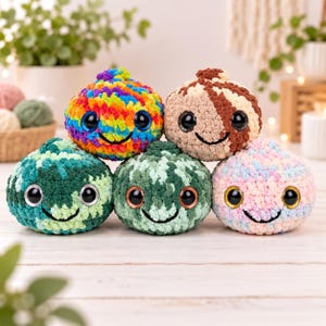 Puede incluir: Cinco juguetes redondos de ganchillo, acolchados, con grandes ojos negros brillantes y sonrisas bordadas. Los juguetes presentan varios colores de hilo, incluyendo un patrón de arcoíris, un remolino marrón y crema, y tres tonos de hilo verde y rosa.