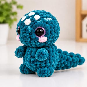 Puede incluir: Un peluche de ganchillo color verde azulado con forma de dinosaurio, cabeza grande y redonda, cuerpo de hilo texturizado y cola larga. Tiene ojos negros grandes con destellos morados, mejillas rosadas y manchas blancas en la parte superior.