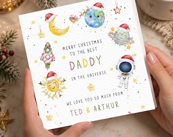 May include: A square greeting card with watercolor space illustrations including a rocket, planets, a sun, and an astronaut, all wearing Santa hats. Text reads: MERRY CHRISTMAS TO THE BEST DADDY IN THE UNIVERSE WE LOVE YOU SO MUCH FROM TED & ARTHUR.