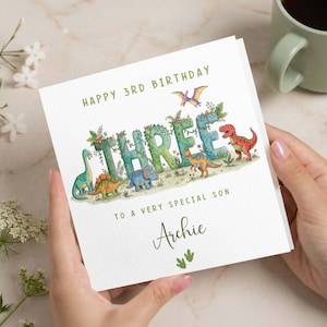 May include: A white birthday card with the words "HAPPY 3RD BIRTHDAY" in green. The word "THREE" is decorated with dinosaurs and foliage. The card reads "TO A VERY SPECIAL SON" and is personalized with the name "Archie".