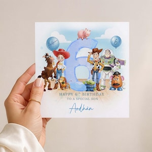 May include: A birthday card with Toy Story characters, including Woody and Buzz Lightyear, and a large blue number 6. The card says "Happy 6th Birthday to a special son" and has a name in blue script. The card is held by a hand.