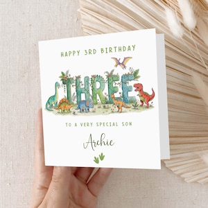 May include: A white birthday card with the text "HAPPY 3RD BIRTHDAY" in green. The word "THREE" is decorated with leaves and dinosaurs. The card reads "TO A VERY SPECIAL SON Archie".