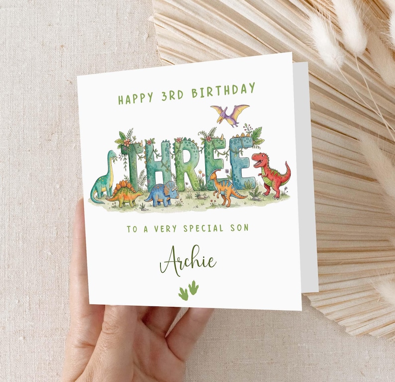 May include: A white birthday card with the text "HAPPY 3RD BIRTHDAY" in green. The word "THREE" is decorated with leaves and dinosaurs. The card reads "TO A VERY SPECIAL SON Archie".