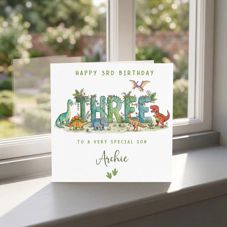 May include: A birthday card featuring the words "HAPPY 3RD BIRTHDAY" and "THREE" in green, adorned with dinosaur illustrations. The card also includes the text "TO A VERY SPECIAL SON" and the name "Archie".