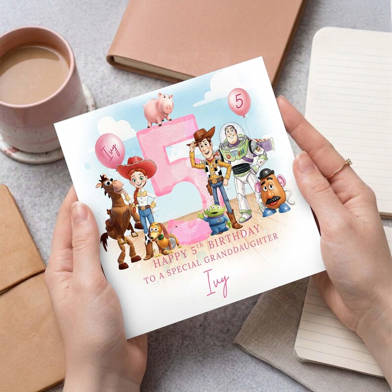 May include: A birthday card with Toy Story characters, a pink number 5, and balloons. The card reads "Happy 5th Birthday to a Special Granddaughter Ivy". The card is held in hands.