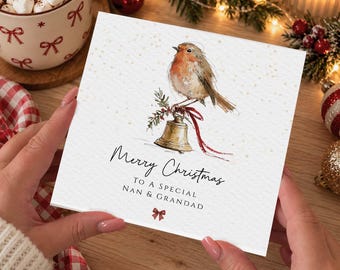 May include: A square white greeting card featuring a watercolor illustration of a robin perched on a gold bell tied with a red ribbon. The text reads Merry Christmas TO A SPECIAL NAN & GRANDAD with a small red bow icon below the text.