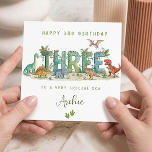 May include: A white birthday card with the text "HAPPY 3RD BIRTHDAY" and "THREE" in large, green, leafy letters. The card features dinosaur illustrations and the name "Archie".