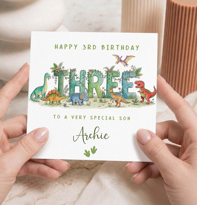 May include: A white birthday card with the text "HAPPY 3RD BIRTHDAY" and "THREE" in large, green, leafy letters. The card features dinosaur illustrations and the name "Archie".
