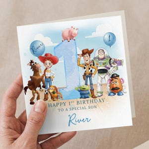 May include: A birthday card with Toy Story characters, a large blue number 1, and a balloon. The card reads "Happy 1st Birthday To A Special Son River". The card is held by a hand.