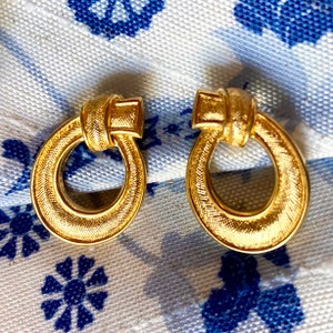 May include: A pair of gold-toned door knocker style earrings. The earrings are round and have a textured surface.