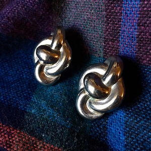 May include: A pair of silver knot-shaped earrings. The earrings are made of a smooth, shiny metal and have a simple, elegant design.