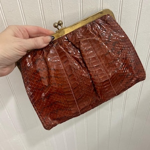 May include: A vintage brown clutch with a reptile skin texture. The rectangular bag has a gold-toned clasp closure. The bag is held against a white background.