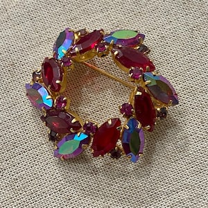 May include: A vintage gold-tone brooch in a circular wreath design. The brooch features red and iridescent marquise-cut rhinestones. Small, round, red rhinestones fill the gaps between the larger stones. The brooch is set on a textured, neutral-coloured surface.