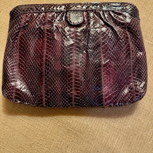 May include: A burgundy clutch with a snakeskin pattern. The rectangular bag has a rounded top and a small, circular clasp. The snakeskin texture is visible across the entire surface of the bag, creating a luxurious look.
