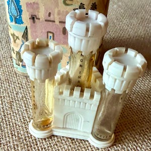 Op de afbeelding: Een vintage parfumset met drie doorzichtige glazen flesjes in een witte, kasteelvormige kunststof basis. Elk flesje is voorzien van een witte, gekanteelde torenkap. De glazen flesjes zijn voorzien van de tekst MONIMAC en LUCIEN.