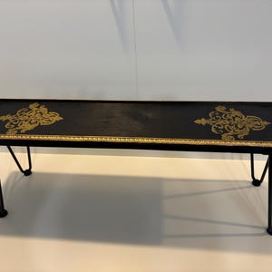May include: A rectangular black coffee table with gold trim and ornate gold designs. The table has black hairpin legs. The table is approximately 48 inches long and 16 inches wide.