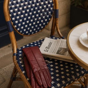 air of burgundy long leather opera gloves with center seam detailing, displayed on a woven café chair beside a newspaper and table.
