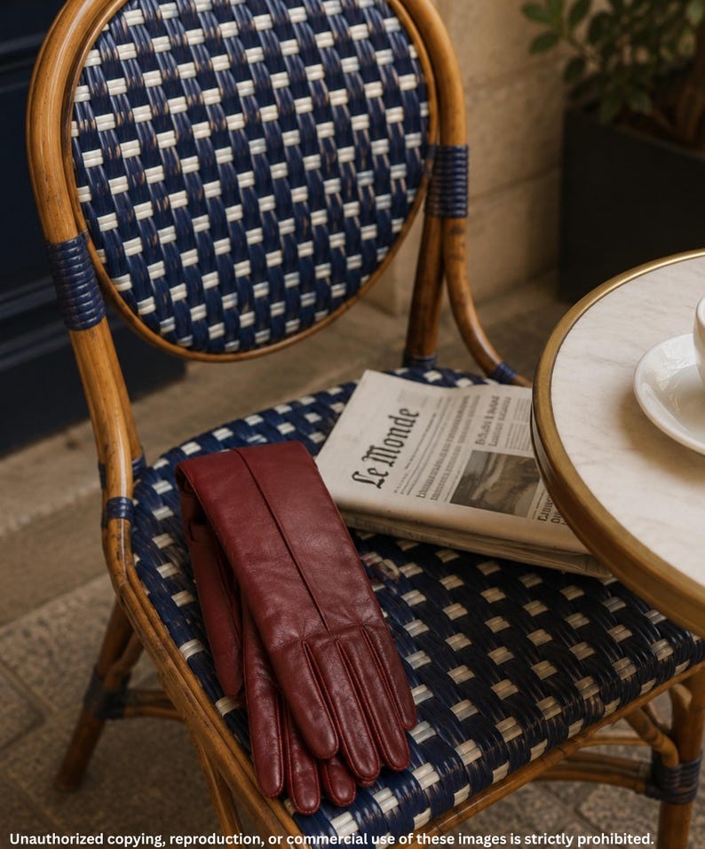 air of burgundy long leather opera gloves with center seam detailing, displayed on a woven café chair beside a newspaper and table.