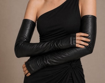 Könnte beinhalten: Schwarze Leder-Opernhandschuhe, die von den Fingerspitzen bis zum Oberarm reichen. Die Handschuhe sind eng anliegend und haben eine glatte Textur. Das Model trägt ein schwarzes One-Shoulder-Kleid.