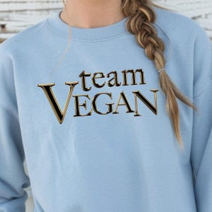 Op de afbeelding: Een lichtblauw sweatshirt met de tekst "team VEGAN" in een goudkleurig en zwart lettertype. Het sweatshirt is effen van kleur en heeft een ronde hals.