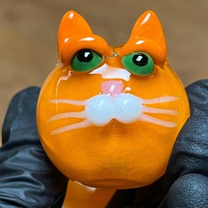 以下が含まれることがあります： 緑色の目、白いひげ、ピンク色の鼻を持つオレンジ色のガラス製の猫の置物です。