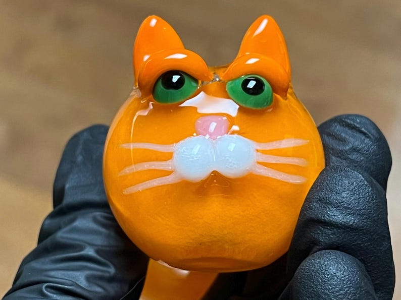 以下が含まれることがあります： 緑色の目、白いひげ、ピンク色の鼻を持つオレンジ色のガラス製の猫の置物です。